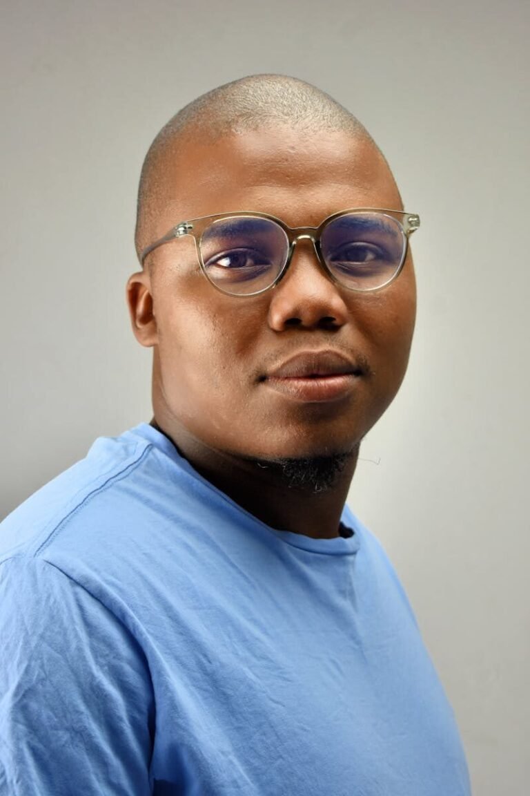 A confident young man wearing glasses and a blue shirt against a neutral background.