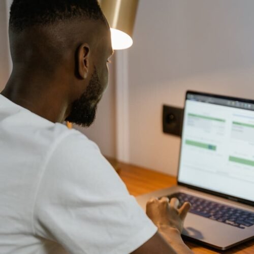 Adult man using laptop at home desk, focused on screen under warm desk lamp light.